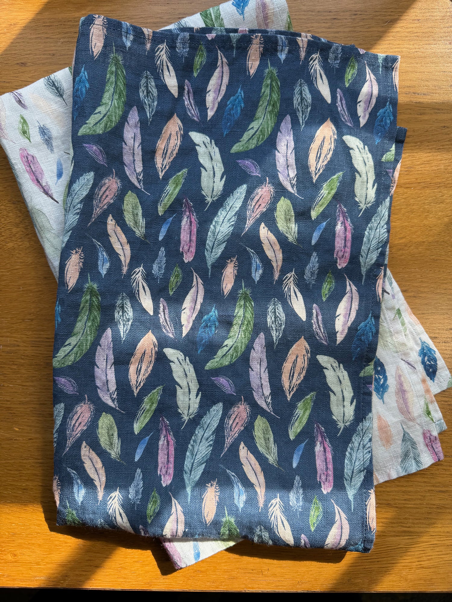 Two folded Tea Towels with feather pattern on a wooden surface